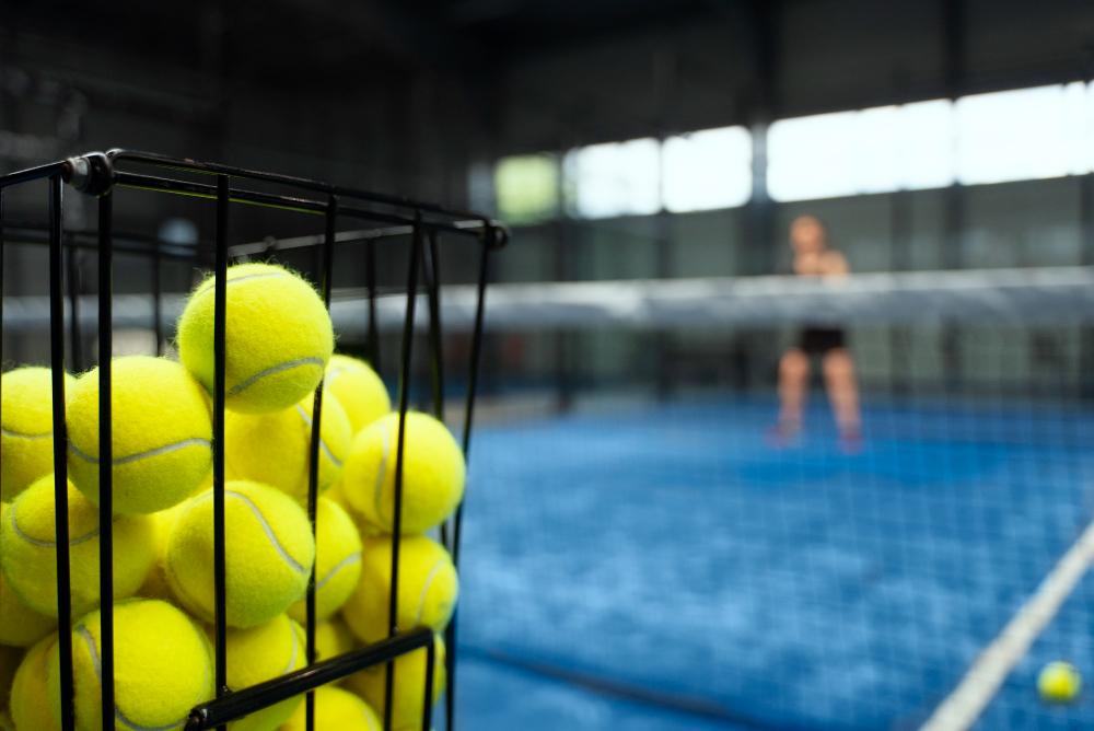 Basket of padel balls on indoor court with player in background, illustrating padel vs pickleball comparison